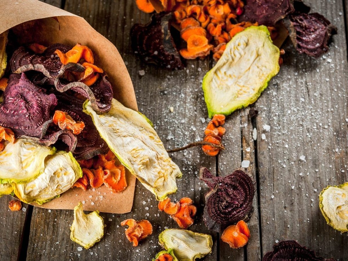 Eine Portion Gemüse-Chips auf einem dunklen Holztisch. Die Chips bestehen aus Zucchini, Möhren und Rote Bete. Auf dem Tisch ist außerdem Salz verteilt.