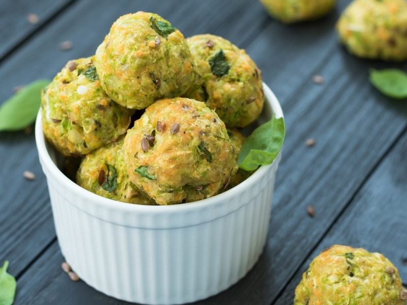 Gemüsebällchen mit Spinat und Parmesan stehen in einer weißen Schüssel auf einem dunklen Holztisch. Die Gemüsebällchen sind angerichtet mit etwas Basilikum und Pinienkernen. Um die Schale herum liegen weitere Gemüsebällchen verteilt.