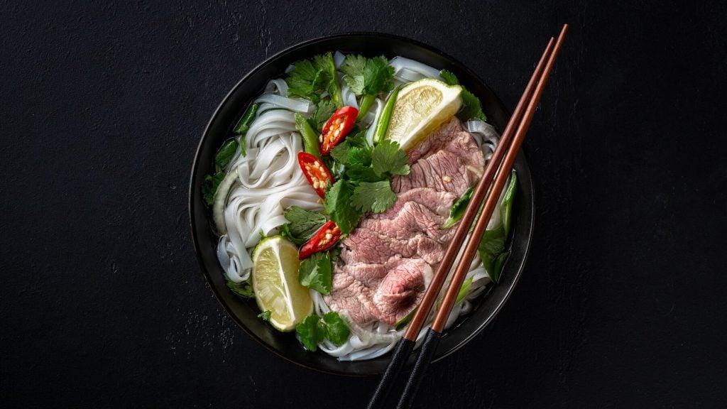 Eine Portion Pho mit Rindfleisch auf einem schwarzen Untergrund. In der Suppe sind außerdem Reisnudeln, Kräuter, Limettenscheiben und Chilischoten. Auf der Schüssel liegen zwei braune Essstäbchen.