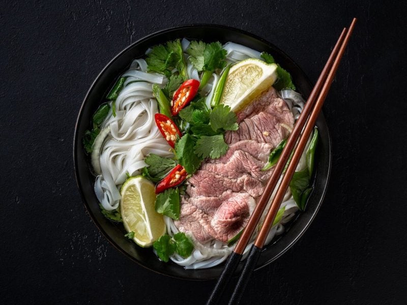 Eine Portion Pho mit Rindfleisch auf einem schwarzen Untergrund. In der Suppe sind außerdem Reisnudeln, Kräuter, Limettenscheiben und Chilischoten. Auf der Schüssel liegen zwei braune Essstäbchen.