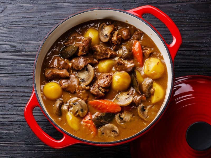 Roter Topf gefüllt mit Pilzgulasch mit Kartoffeln und Gemüse auf dunklem Holzuntergrund.