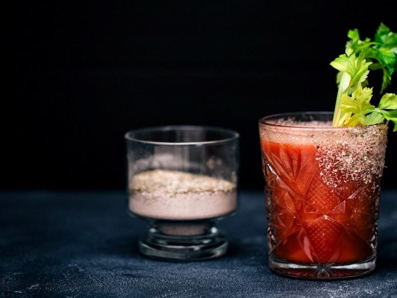 Ein Virgin Bloody Mary in einem Glas mit Selerie. Das Glas ist angerichtet mit Salz und Pfeffer. In einem Glas daneben steht eine Mischung aus Salz und Pfeffer.