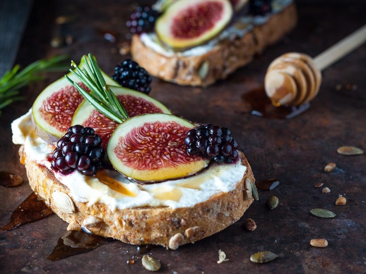 Vollkornbrot mit Feigen, Ricotta und Honig auf einem dunklen Untergrund. Im Hintergrund liegt ein Honiglöffel. Auf dem Untergrund verteilt sind Thymian, Körner und Honig.