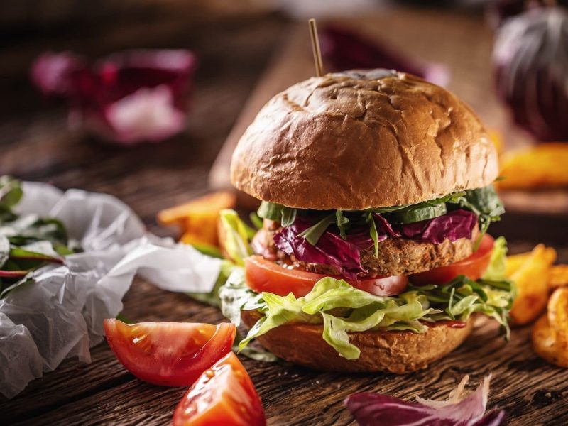 Bellas Gemüse-Burger auf einem Holztisch. Um den Burger herum liegen Pommes, Salat und Tomaten. Der Burger wird mit einem Holzstab zusammen gehalten