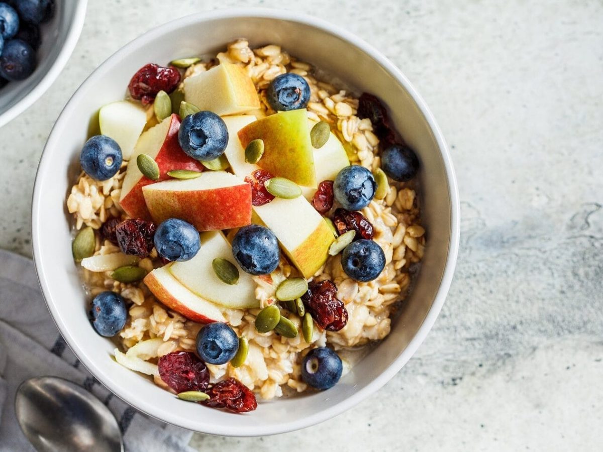 Apfelschnitze, Blaubeeren, getrocknete Cranberries, Pistazien, Walnüsse und Haferflocken toppen den Joghurt in einer Schüssel und ergeben das Bircher Müsli.