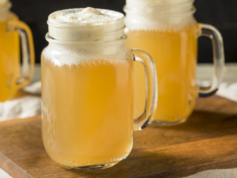 Zwei gläserne Krüge mit Butter-Bier stehen auf einem Holzbrett auf einem Tisch mit einer beigen Tischdecke. Im Hintergrund steht ein weiteres Butter-Bier. Die Biere sind angerichtet mit Sahne.