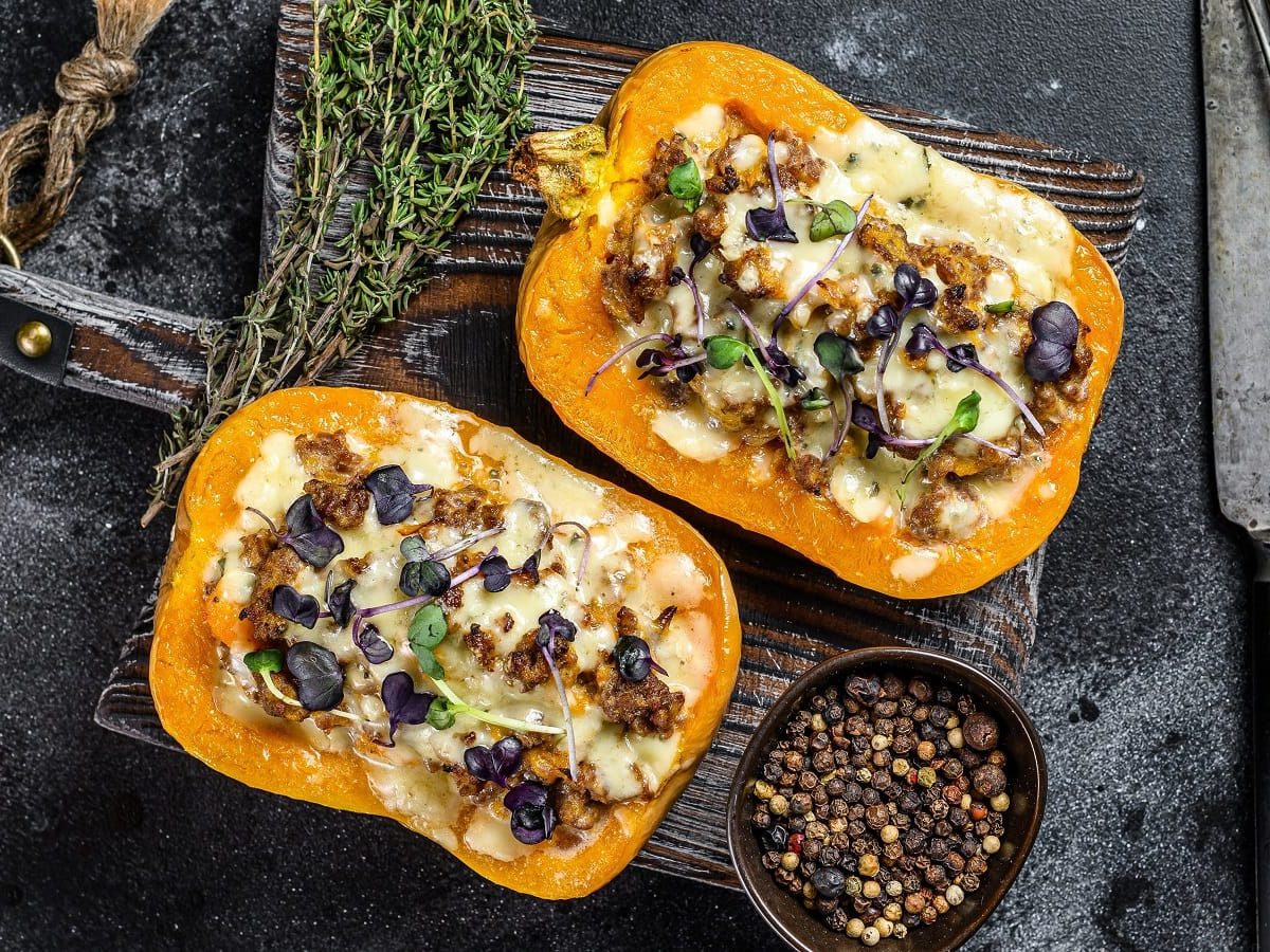 Zwei Hälften Butternutkürbis mit Reis, Hackfleisch unf Pfifferlingen gefüllt, Käse überbacken und frischen Kräutern garniert, auf einem Holzbrett neben Thymian Stengeln und einer Schüssel Pfefferkörner, vor dunklem Hintergrund von oben.