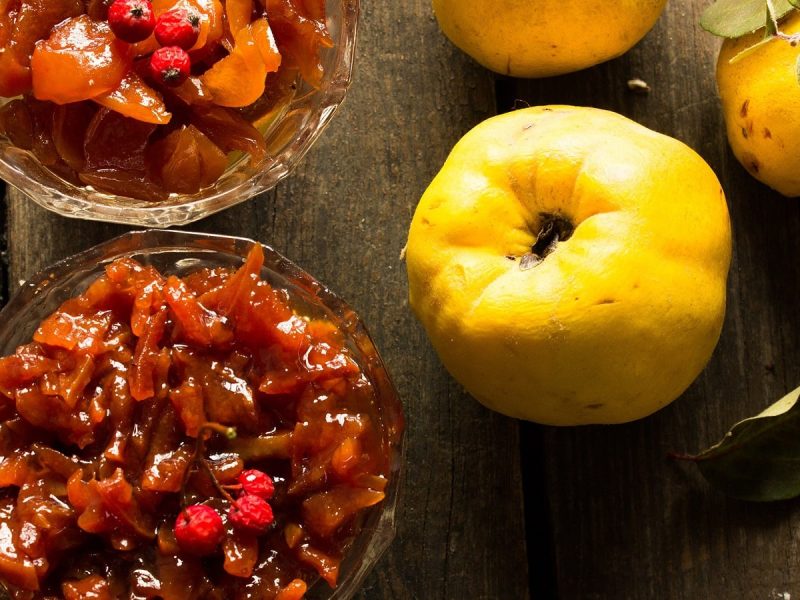 Chutney mit Quitten und Äpfeln in zwei Glasschüsseln angerichtet neben frischen Quitten vor einem Holz-Hintergrund von oben.