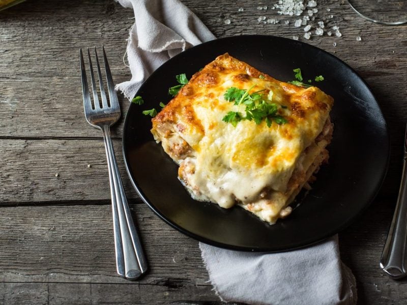 Ein Stück Lasagne auf einem schwarzen Teller angerichtet mit Besteck. Im Hintergrund die Auflaufform und ein Glas Wein.