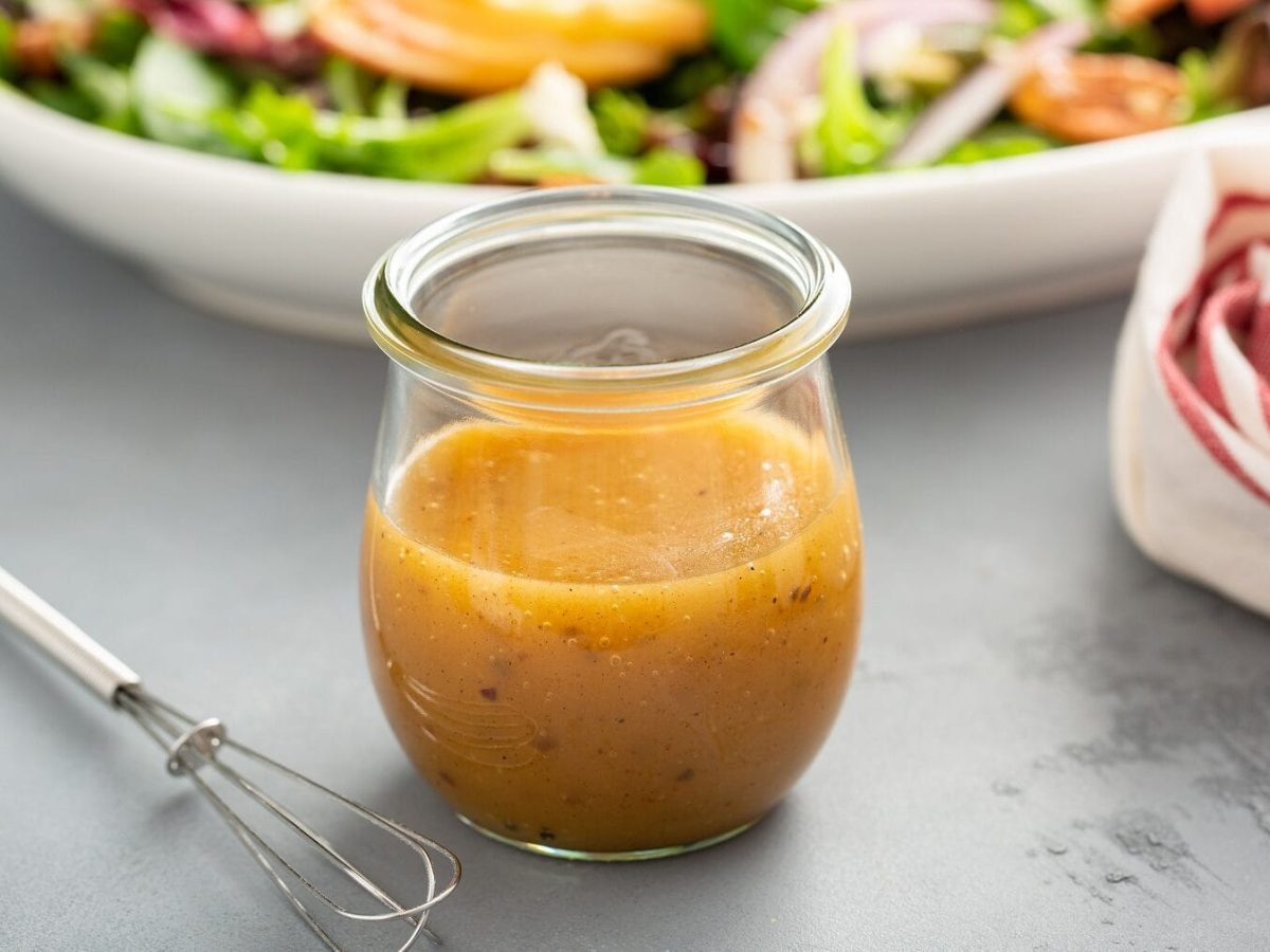 EIn Glas mit Dressing steht auf einem grauen Tisch. Neben dem Glas liegt ein kleiner Rührbesen, im Hintergrund steht eine Schale mit Salat. Am rechten Bildrand befindet sich ein weißes Handtuch mit roten Streifen. Die ultimative Dressingformel.