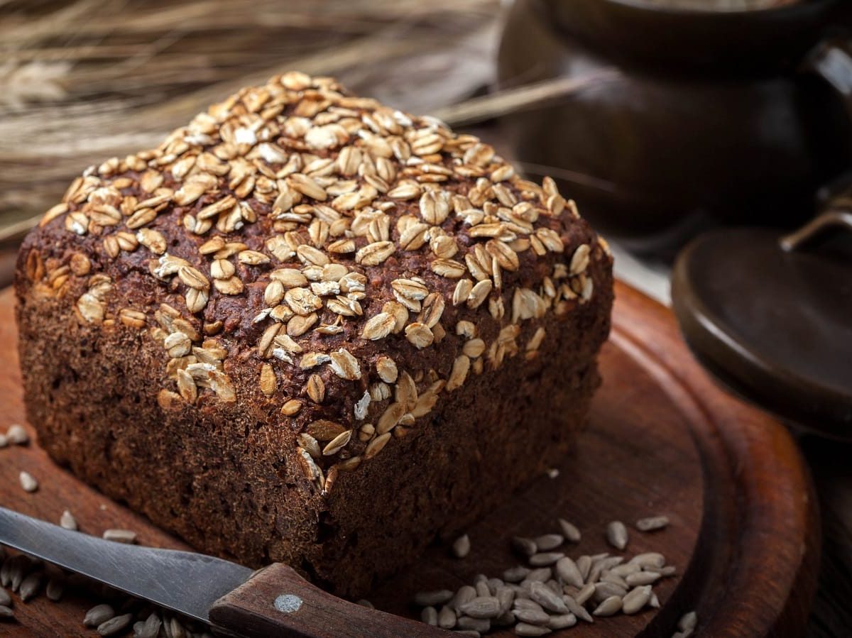 Ein halbes Laib Dreikornbrot in kastiger Form mit Haferflocken und Kürbiskernen bestreut auf einem hölzernen Schneidebrett neben Ehren und einem Tonkrug.