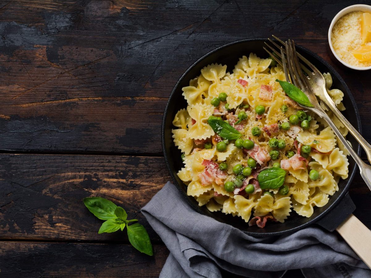 Auf einem Holztisch steht eine Pfanne. In der Pfanne sind Nudel, Schinken, Speck und Basilikum. Daneben steht ein Schälchen mit Parmesan.