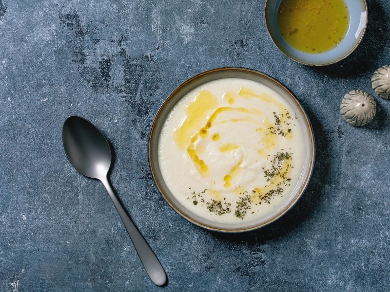 Eine Schüssel mit einer Portion weißer Fenchel-Creme-Suppe garniert mit Gewürzen und Olivenöl neben salz und Pfefferstreuer, einer kleinen Schüssel Olivenöl sowie einem Löffel, vor grauem Hintergrund von oben.