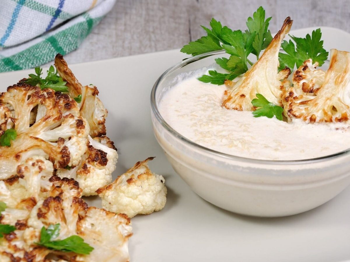 Eine Portion gebackener Blumenkohl mit Tahini-Sauce steht auf einem weißen Tablett auf einem grauen Untergrund. Im Hintergrund liegt ein Geschirrtuch. Der Blumenkohl ist angerichtet mit Petersilie. Die Tahini-Soße befindet sich in einer kleinen Glasschüssel.