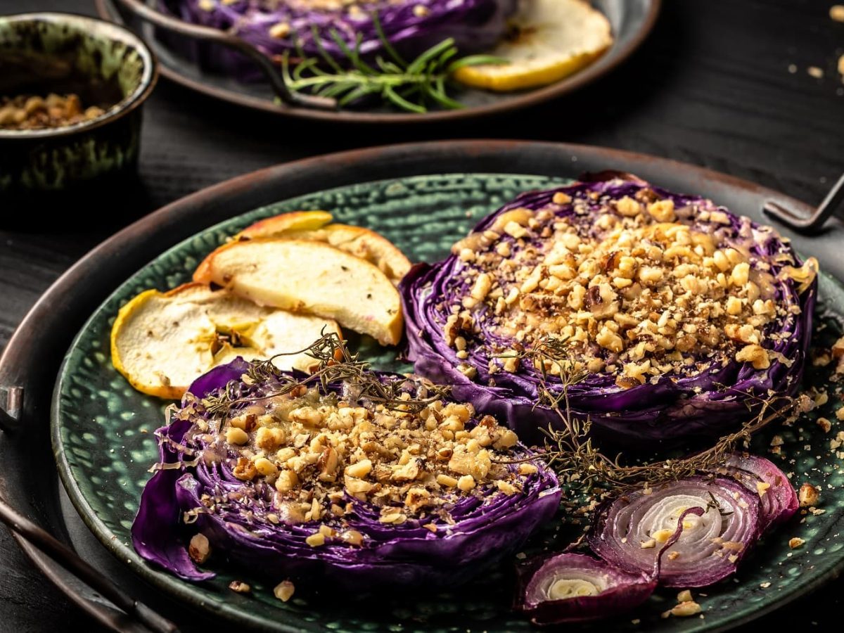 Gebackener Rotkohl mit Nüssen und Äpfeln auf einem gemusterten Teller auf einem Tablett. Der Rotkohl ist mit Zwiebelscheiben und gebackenem Apfel angerichtet.