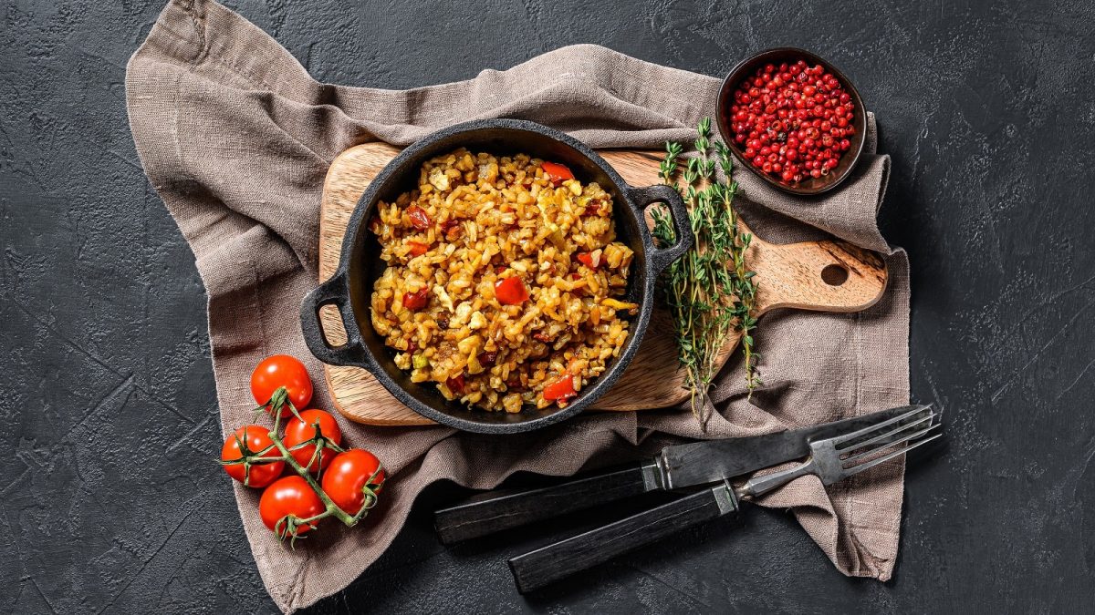 Eine schwarze Pfanne gefüllt mit gebratenem Reis mit Tomaten und Cheddar auf einem Holzbrett neben Thymian-Zweigen einer Schüssel Pfeffer, frischen Tomaten und Besteck auf einem grau-braunen Küchentuch vor einem schwarzen Hintergrund, von oben.