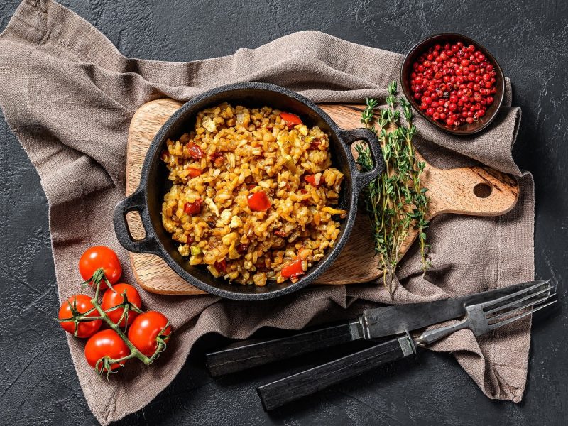 Eine schwarze Pfanne gefüllt mit gebratenem Reis mit Tomaten und Cheddar auf einem Holzbrett neben Thymian-Zweigen einer Schüssel Pfeffer, frischen Tomaten und Besteck auf einem grau-braunen Küchentuch vor einem schwarzen Hintergrund, von oben.