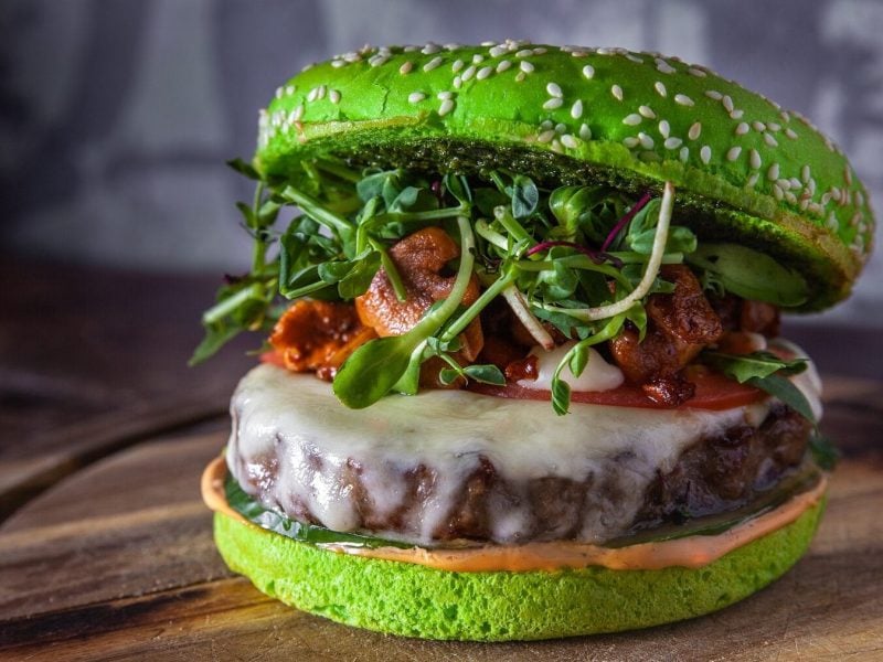 Ein grüner Burger mit Sprossen und Pilzen steht auf einem braunen Holzbrett auf einem hölzernen Untergrund. Der Hintergrund ist grau und gemustert. Auf dem Burgerbrötchen befindet sich Sesam. Der Burger ist außerdem belegt mit einem Burgerpatty, Hackfleisch, einer Tomatenscheibe, Gurke und Soße.