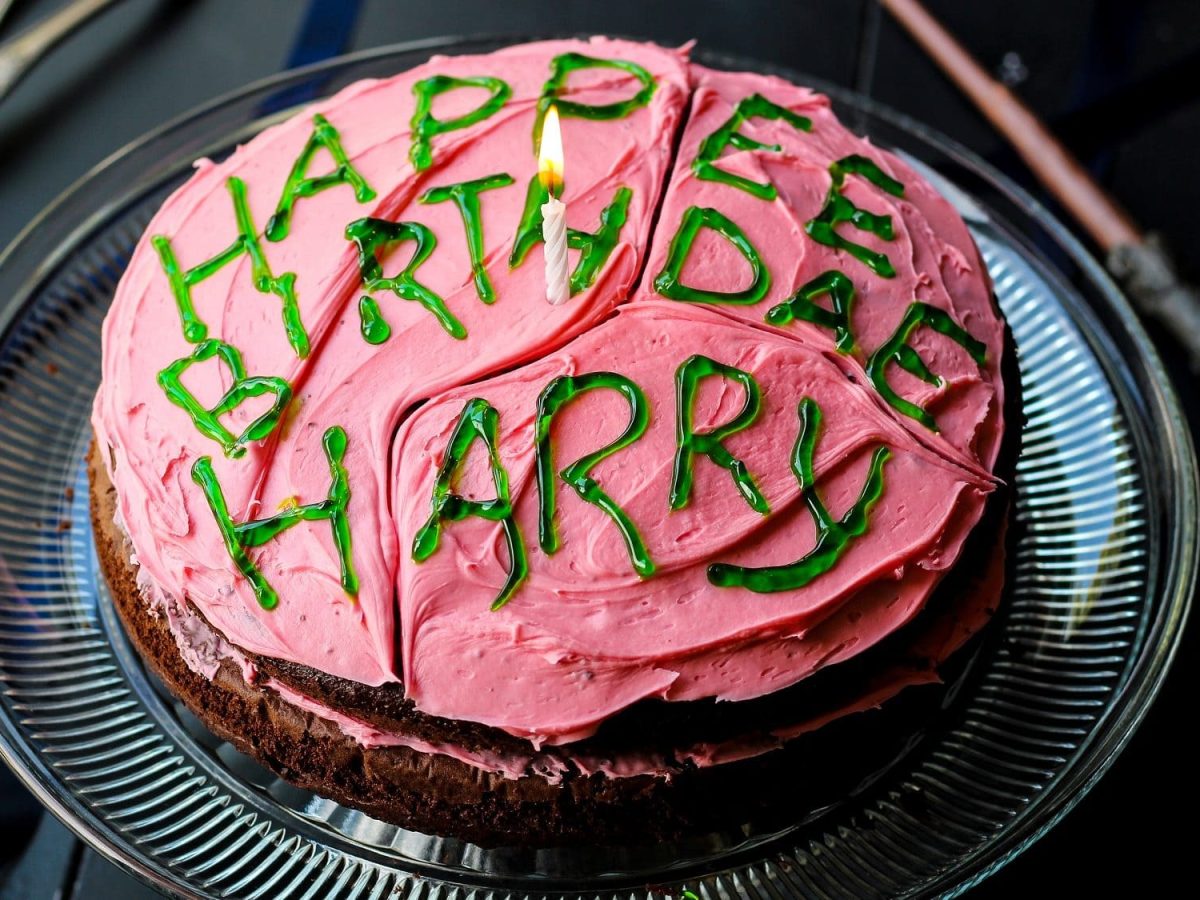 Harry Potters Geburtstagskuchen mit Schokoteig, pinker Buttercreme und grüner Zuckerschrift auf einem Glasteller auf einem schwarzen Tisch. Im Hintergrund liegt ein Zauberstab.