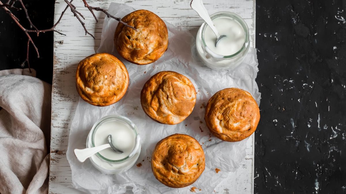 Fünf Kürbis-Frischkäse-Muffins auf einem weißen Backpapier zusammen mit zwei Gläsern Frischkäse angeordnet, vor einem schwar-weißen Hintergrund, von oben.