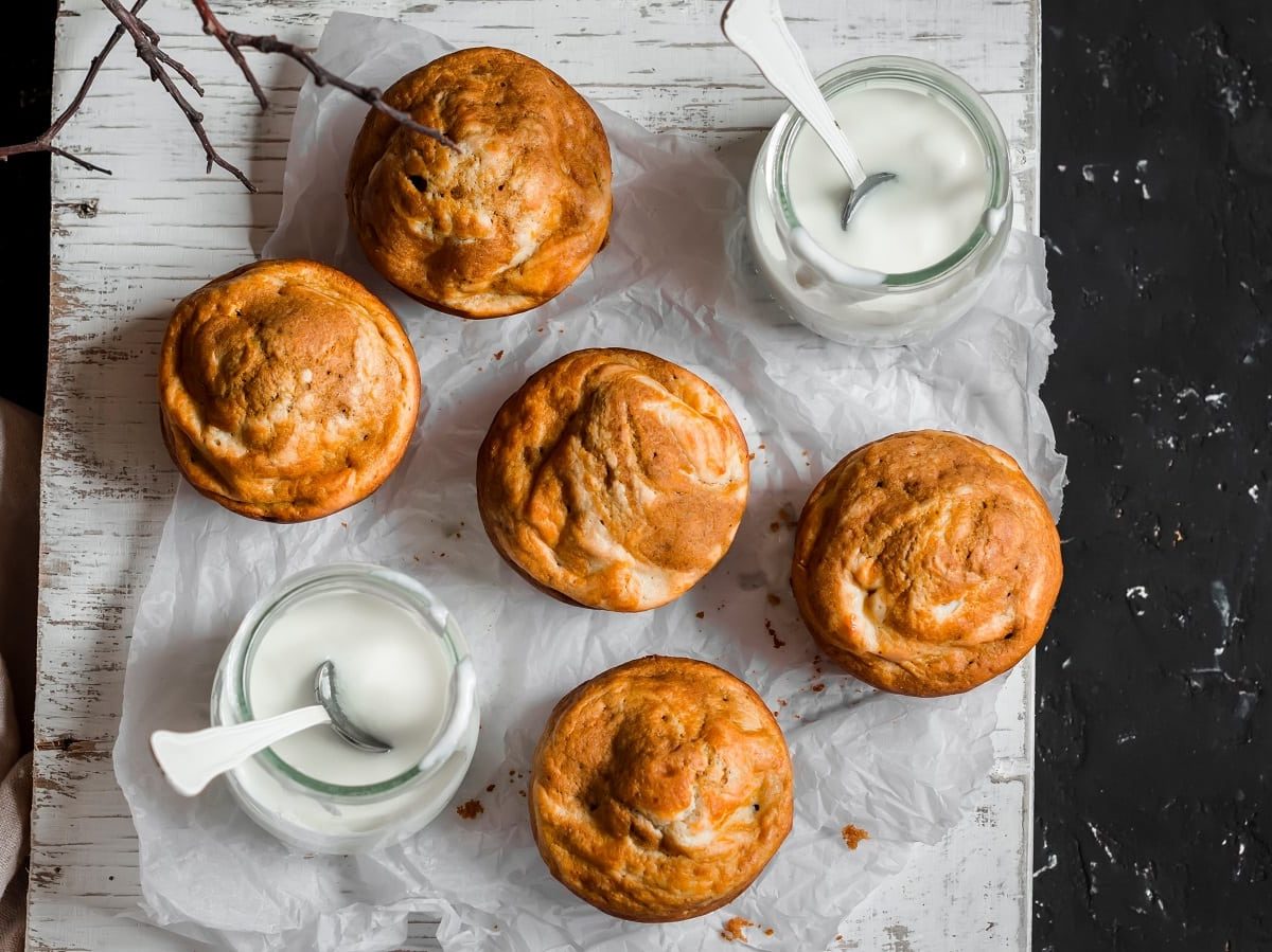 Fünf Kürbis-Frischkäse-Muffins auf einem weißen Backpapier zusammen mit zwei Gläsern Frischkäse angeordnet, vor einem schwar-weißen Hintergrund, von oben.