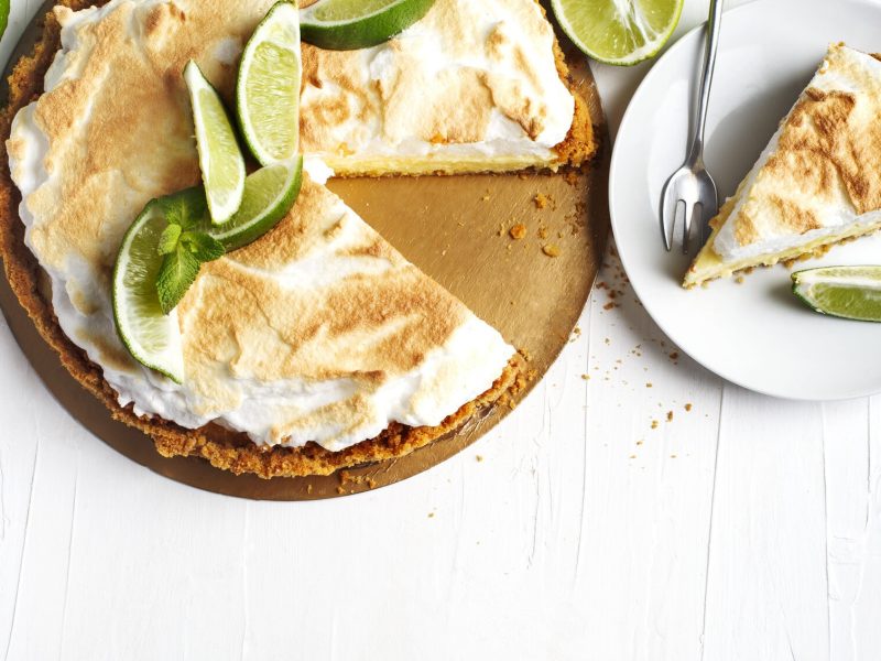 Eine angeschnittene Limetten-Tarte in der Draufsicht, verziert mit Limettenscheiben und Baiserhaube. Daneben steht ein Teller mit einem Stück Tarte.