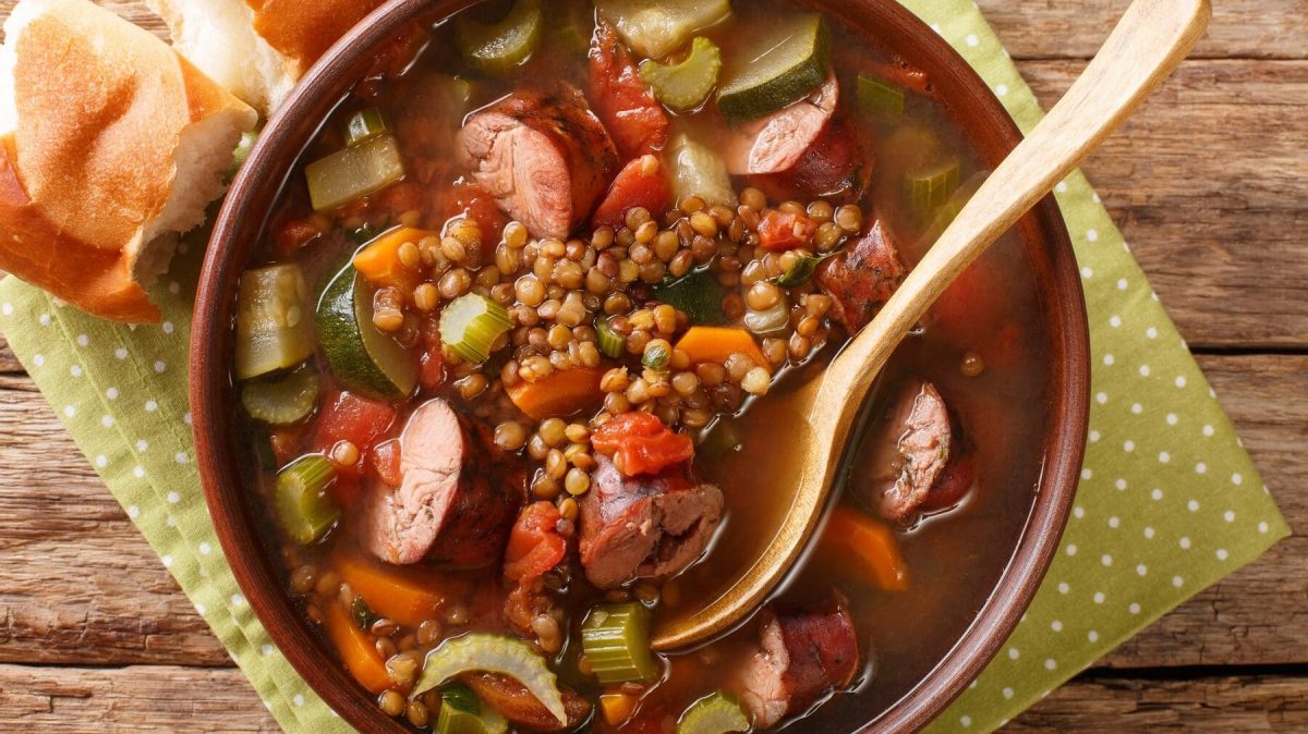 Eine Portion Linsensuppe mit Mettwürstchen in einer braunen Schüssel. Die Schüssle stehta auf einem gepunkteten Geschirtuch auf einem Holztisch. In dem Eintopf steckt ein Löffel aus Holz. Neben der Schüssel liegt ein Stück Baguette.