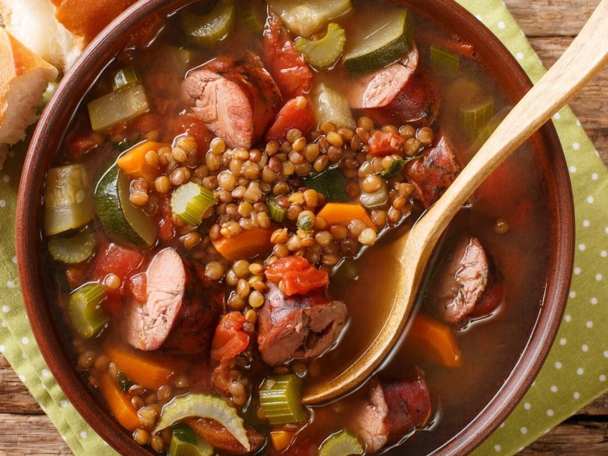 Eine Portion Linsensuppe mit Mettwürstchen in einer braunen Schüssel. Die Schüssle stehta auf einem gepunkteten Geschirtuch auf einem Holztisch. In dem Eintopf steckt ein Löffel aus Holz. Neben der Schüssel liegt ein Stück Baguette.