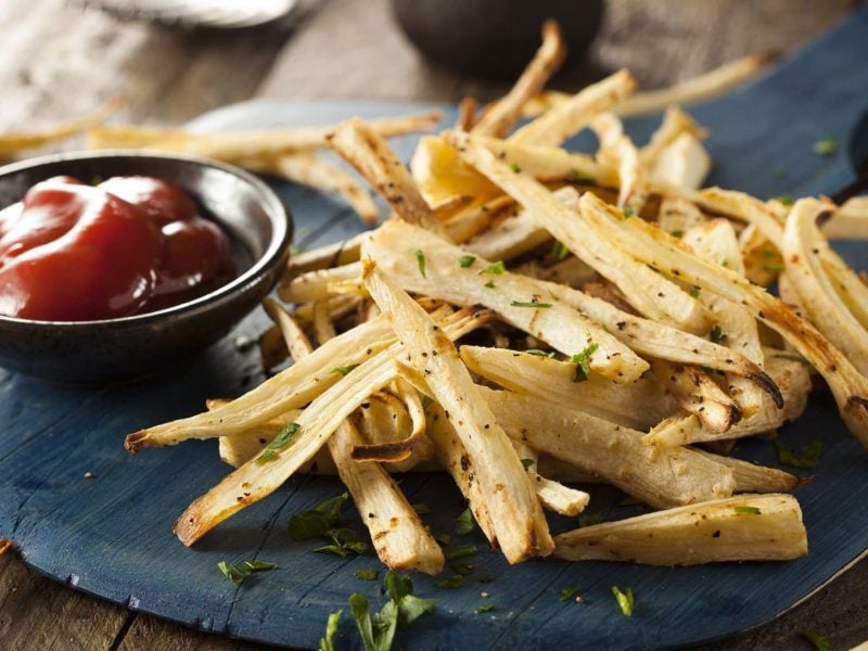 auf einem dunklen Holzbrett liegen Pommes aus Petersilienwurzeln. Danebenn stehte eine kleine Schale mit Ketchup