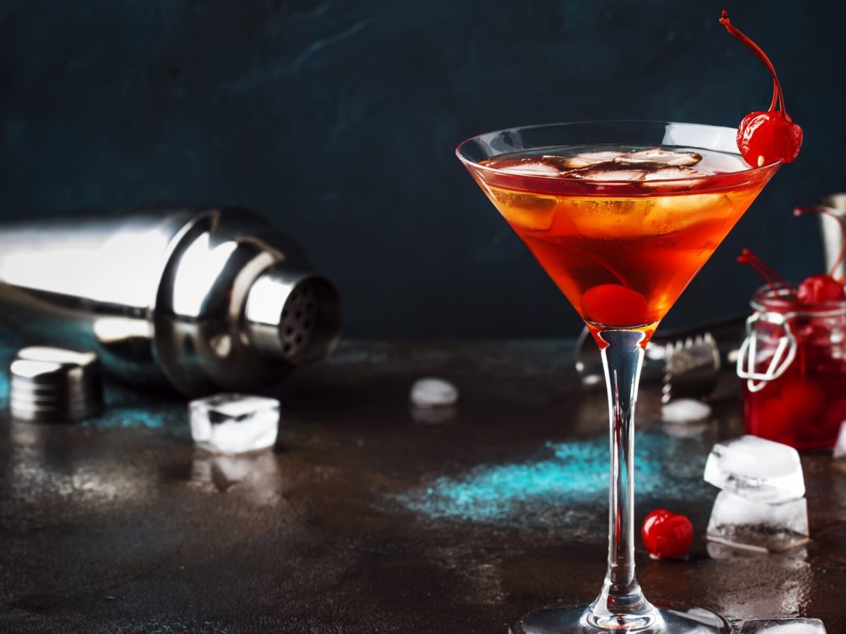 Ein Manhattan-Cocktail in einem Martiniglas, dekoriert mit einer Cocktailkirsche. Im Hintergrund ein umgekippter Cocktail-Shaker und verstreut die Zutaten.