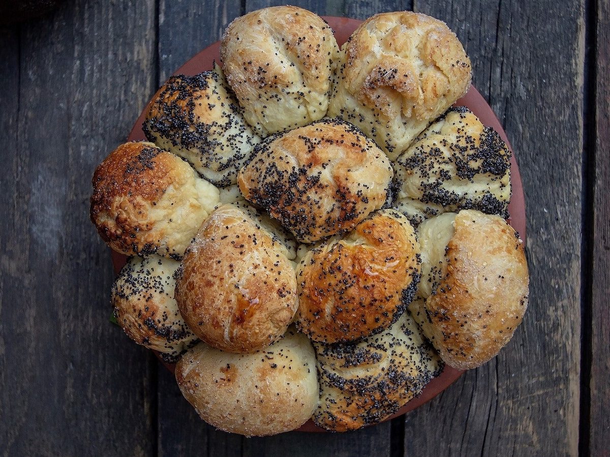 Ein Teller mit einem Monkey-Bread: Kleine Teigkugeln mit Mohn und Salz umhüllt zusammen gebacken. ein einziges großes Brot bildend, vor einem dunklen Holz-Hintergund, von oben.