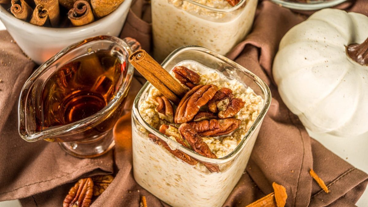 Overnight-Oats mit Ahornsirup und Nüssen auf einem braunen Tuch. Die Oats sind angerichtet mit Pekanüssen und einer Zimtstange. Um die Schale herum stehen ein Zierkürbis, Ahornsirup und Zimtstangen. Auf dem Tuch luegen weitere Pekanüsse.