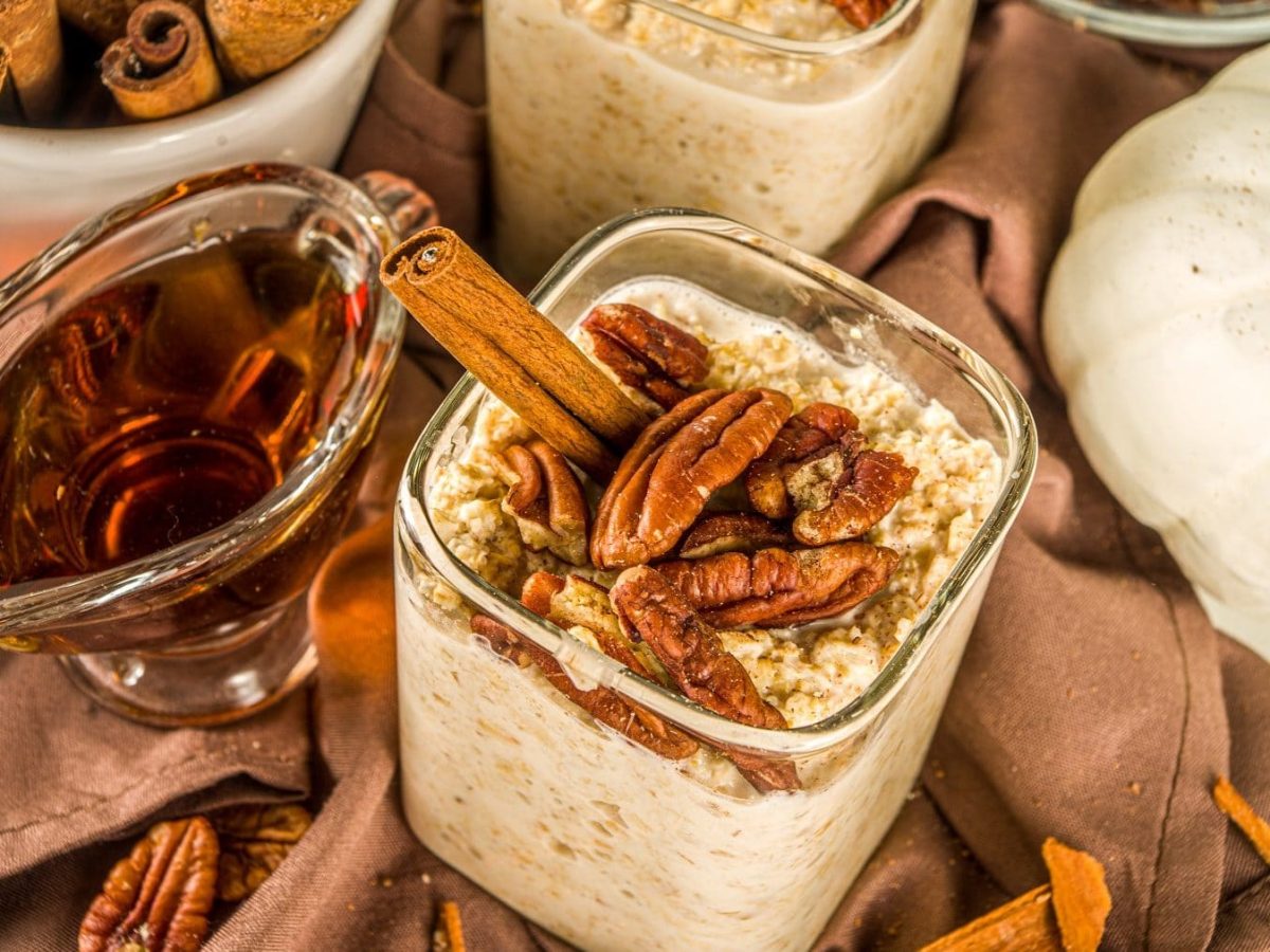 Overnight-Oats mit Ahornsirup und Nüssen auf einem braunen Tuch. Die Oats sind angerichtet mit Pekanüssen und einer Zimtstange. Um die Schale herum stehen ein Zierkürbis, Ahornsirup und Zimtstangen. Auf dem Tuch luegen weitere Pekanüsse.