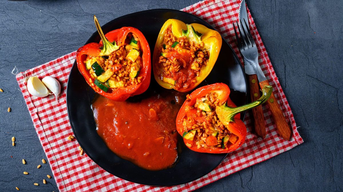 Auf einem dunklen Tisch liegt ein rot-weiß-karierte Tuch. Auf diesem steht ein teller, auf dem zwei rote und eine gelbe Paprika-Hälfte zu sehen ist. Diese sind mit Gemüse gefüllt.. Da gericht heißt: Paprikaschoten mit Gemüse-Bulgur-Füllung