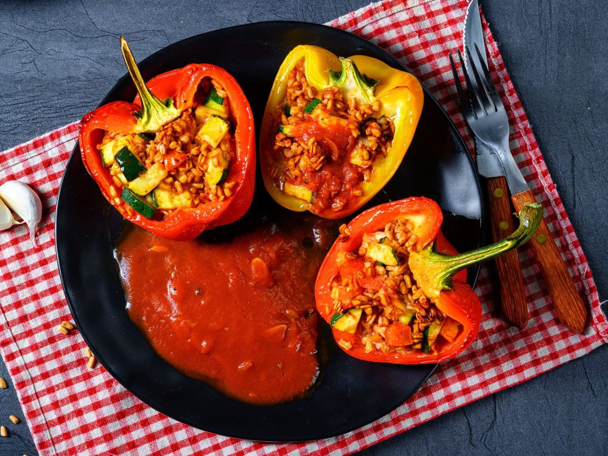 Auf einem dunklen Tisch liegt ein rot-weiß-karierte Tuch. Auf diesem steht ein teller, auf dem zwei rote und eine gelbe Paprika-Hälfte zu sehen ist. Diese sind mit Gemüse gefüllt.. Da gericht heißt: Paprikaschoten mit Gemüse-Bulgur-Füllung