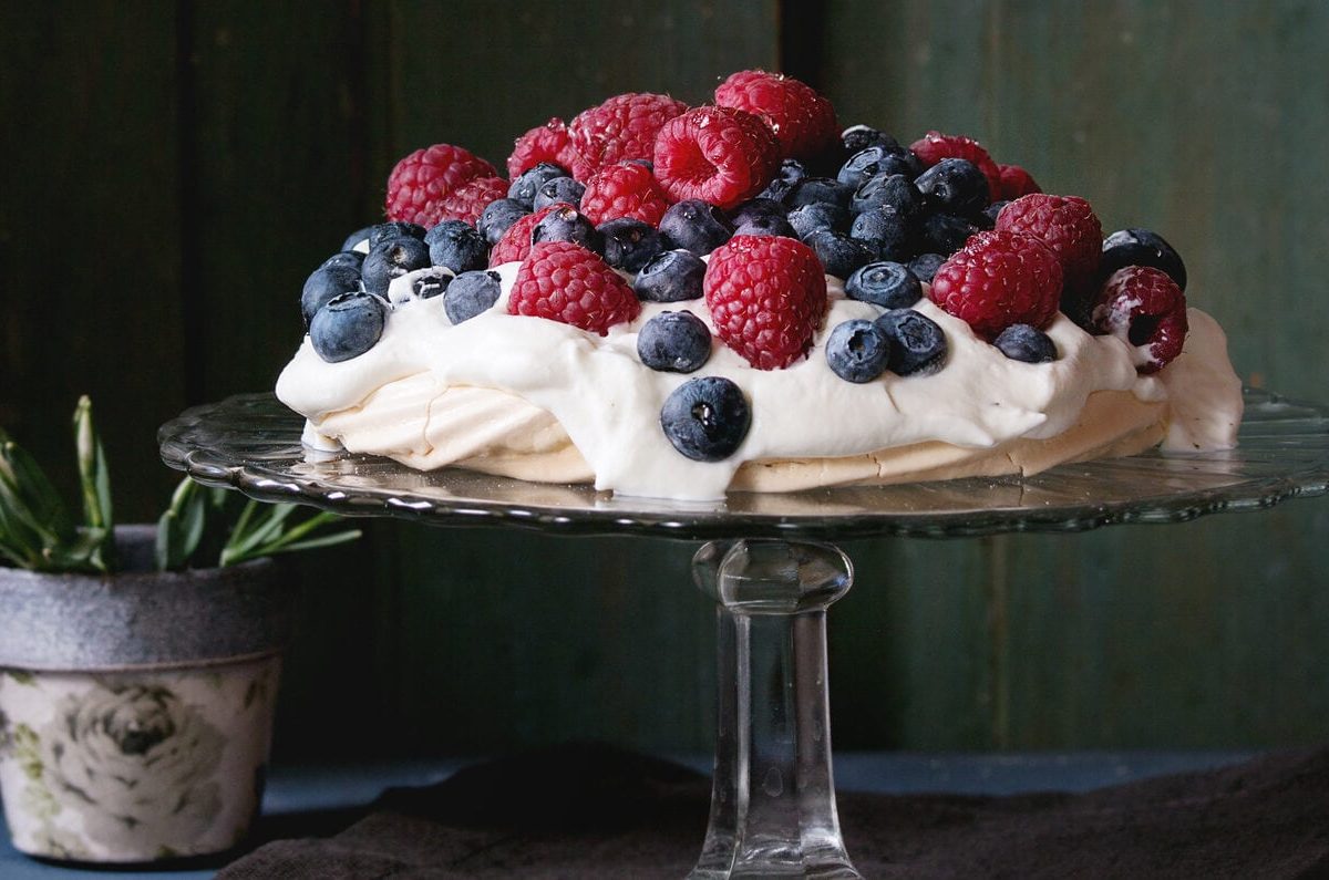 Auf einem Tortenständer steht eine Pavlova mit Beeren vor einem dunkelgrünen Hintergrund.