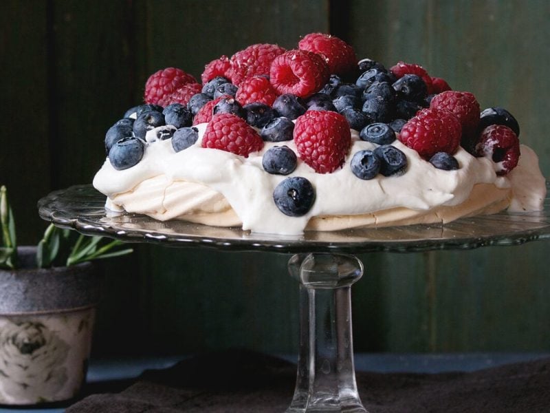 Auf einem Tortenständer steht eine Pavlova mit Beeren vor einem dunkelgrünen Hintergrund.