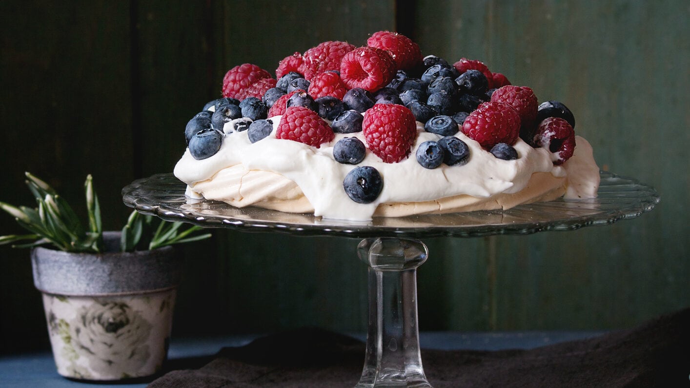 Auf einem Tortenständer steht eine Pavlova mit Beeren vor einem dunkelgrünen Hintergrund.