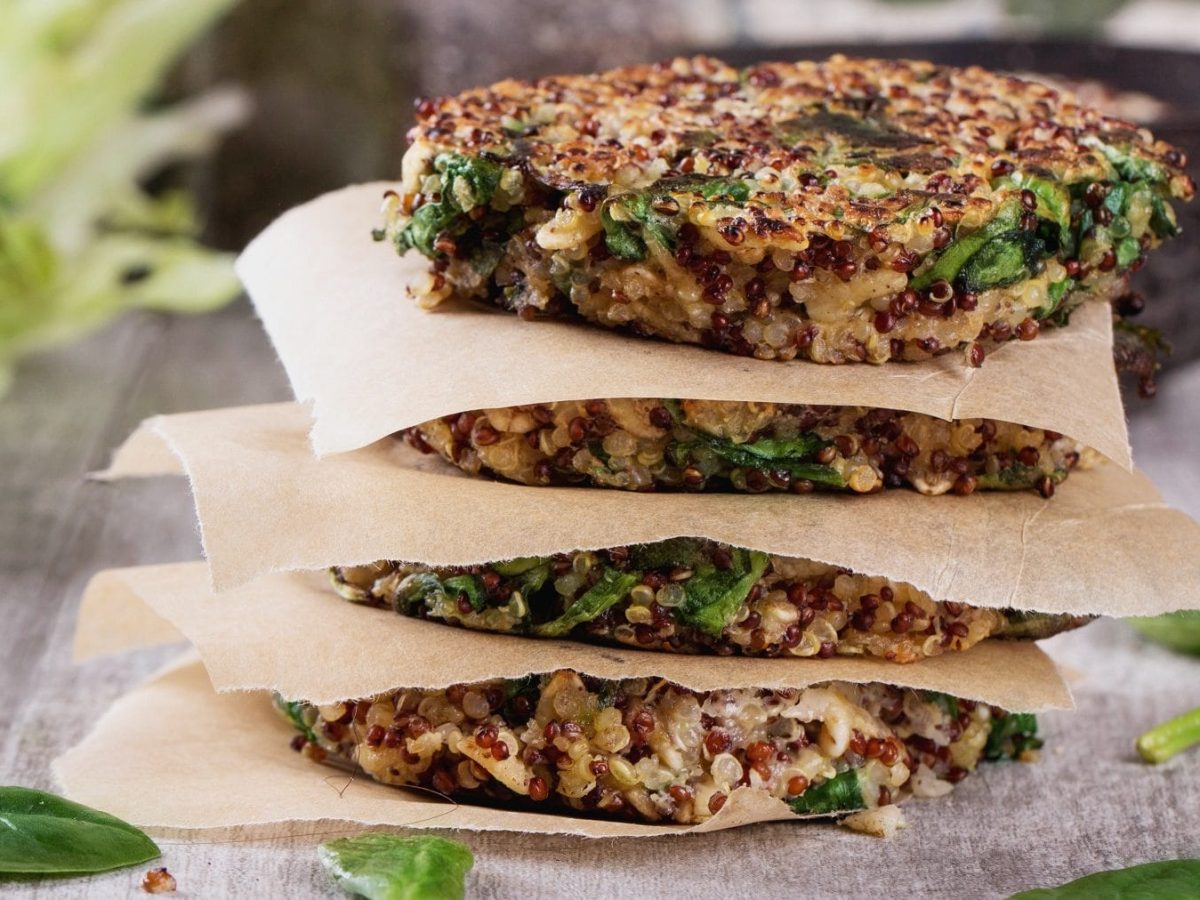 Drei geschichterte Quinoa-Burger-Patties auf einem Untergrund. Zwischen den Patties liegen Backpapierzuschnitte, damit sie nicht aneinander haften. Um die Patties herum liegen Spinatblätter.