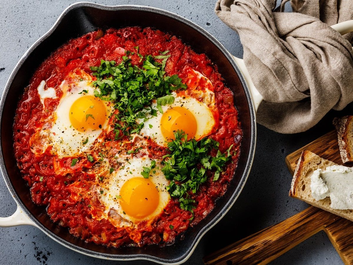 Eine große Pfanne mit Shakshuka mit drei Eiern und viel Koriander und Petersilie garniert neben einem Holzbrett mit mit Frischkäse bestrichenem Brot vor einem grauem Hintergrund, von oben.