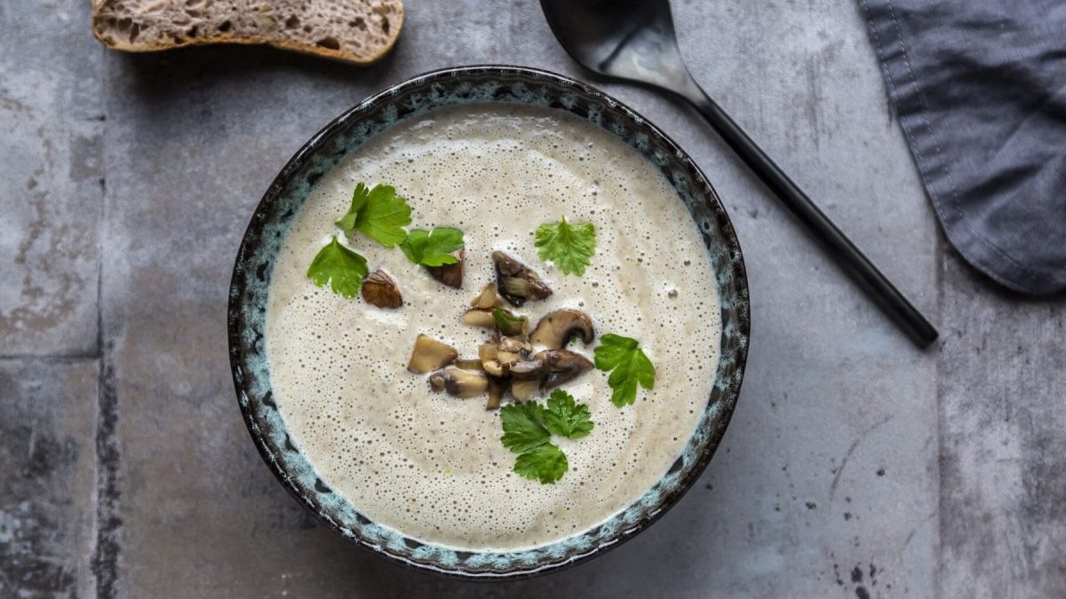 Eine Portion Steckrübensuppe mit Champignons in einer gemusterten Schüssel steht auf einem Steintisch. Neben der Schüssel liegt ein Löffel, am oberen Bildrand liegt eine Scheibe Brot.