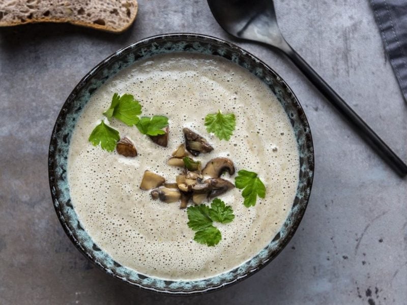 Eine Portion Steckrübensuppe mit Champignons in einer gemusterten Schüssel steht auf einem Steintisch. Neben der Schüssel liegt ein Löffel, am oberen Bildrand liegt eine Scheibe Brot.