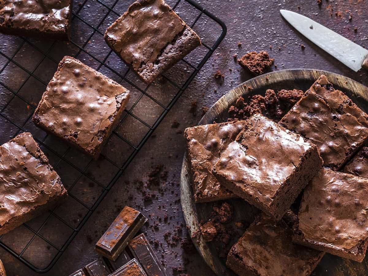 Ein Kuchengitter und rundes Holzbrett mit Süßkartoffel-Brownies in der Draufsicht. Daneben liegen ein Messer und Schokolade.