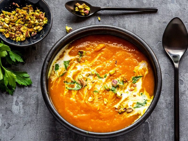 Eine Schüssel mit Süßkartoffelsuppe mit Chili und Pistazien gefüllt und Koriander garniert neben einem großen Lössel, einer kleinen Schale mit gehackten Pistazien und einem kleinen Löffel mit gehackten Pistazien vor einem grauen Hintergrund, von oben.