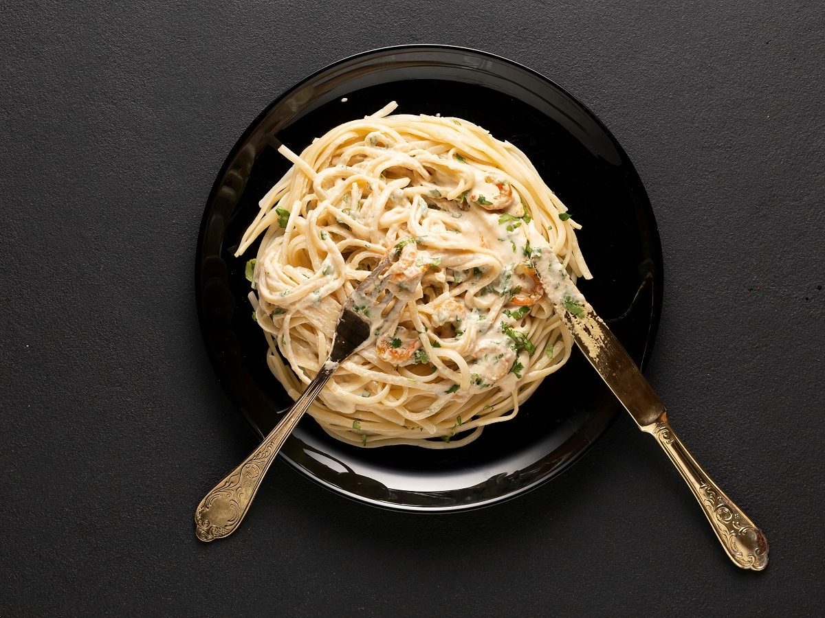 Ein schwarzer Teller mit einer Portion veganer Carbonara mit Pilzen und Messer und Gabel vor einem dunklen Hintergrund von oben.