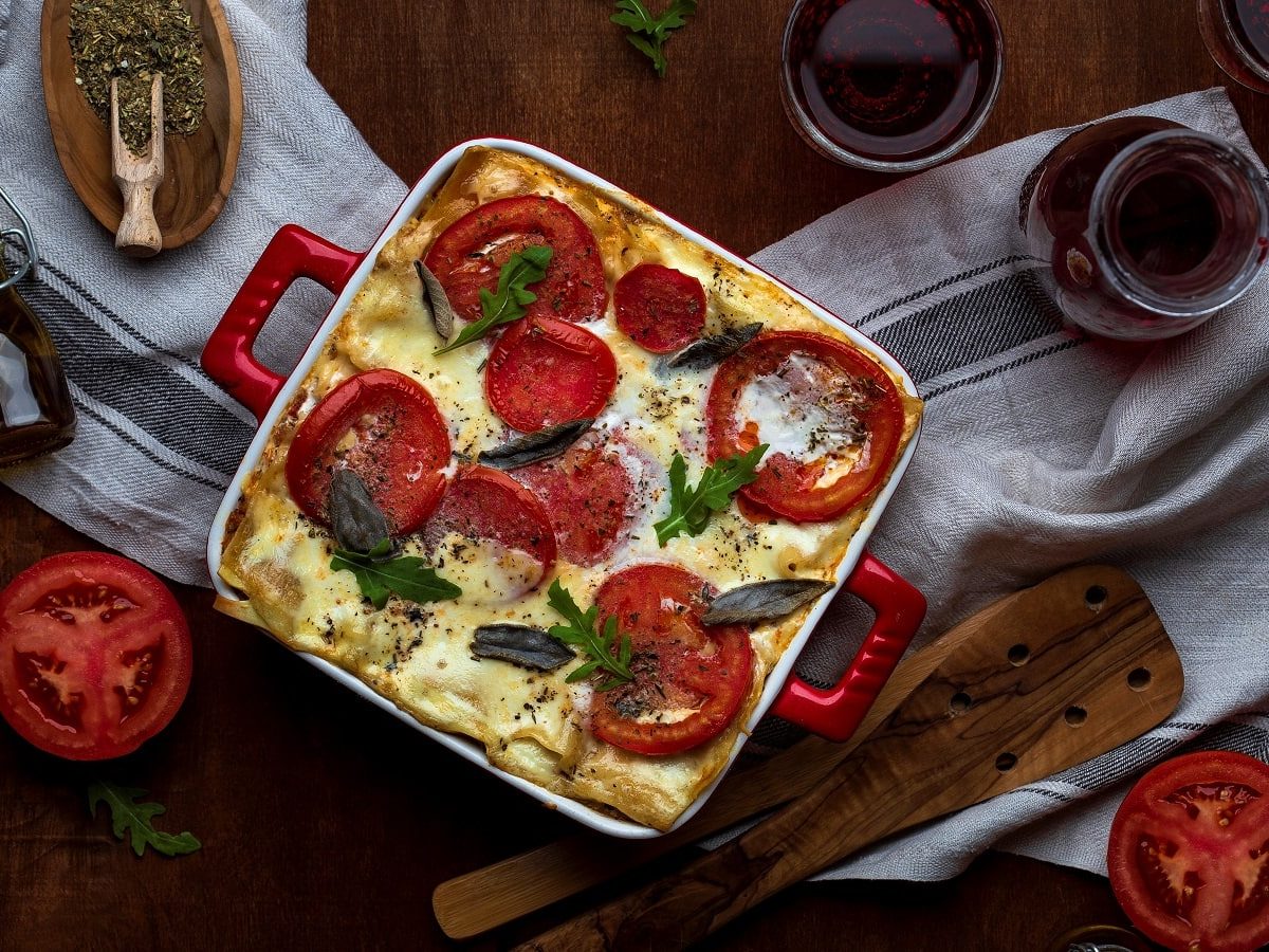 Eine Auflaufform mit veganer Lasagne, die mit Tomaten, veganem Käse, Basilikum und Rucola garniert ist neben einer Karaffe Wein, zwei gefüllten Gläsern, Gewürzen, frischen Tomaten und einem Holzheber, auf einem Küchentuch vor einem dunklen Holzhintergrund, von oben.