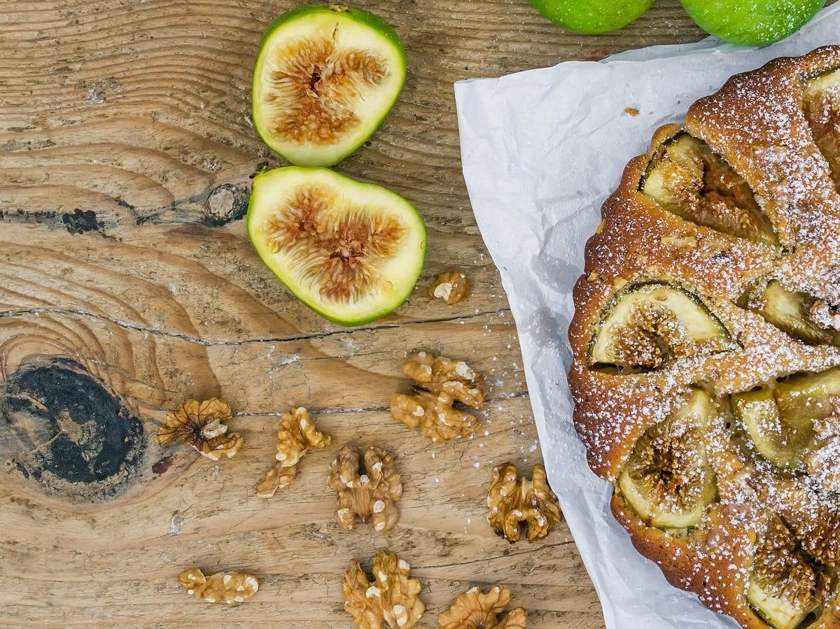 Ein ganzer Walnuss-Feigenkuchen mit Puderzucker bestäubt auf einem weißen Backpapier neben aufgeschnittenen frischen Feigen und Walnusskernen vor einem Holzhintergrund, von oben.