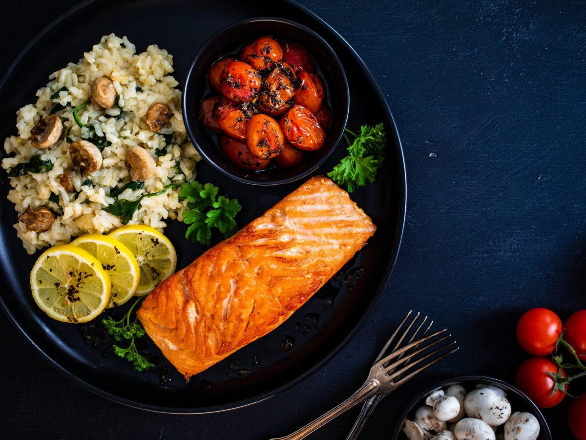 auf einem dunklen Untergrund steht ein dunkler Teller. Auf dem sieht man ein Zitronen-Risotto-mit-Lachs. Außerdem sind Tomaten und Pilze zu sehen.