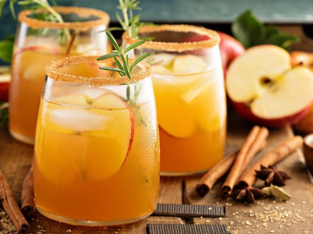 Drei Gläser Apple-Cider-Cocktail mit Zimt und Zucker am Rand. Die Cocktails sind angerichtet mit Eis, Apfel und Gewürzen. Umdie Gläser herum liegen herbstliche Gewürze, Zimtstangen, Zimt und Zucker. Im Hintergrund liegen Äpfel. Die Gläser stehen auf einem braunen Holzbrett.