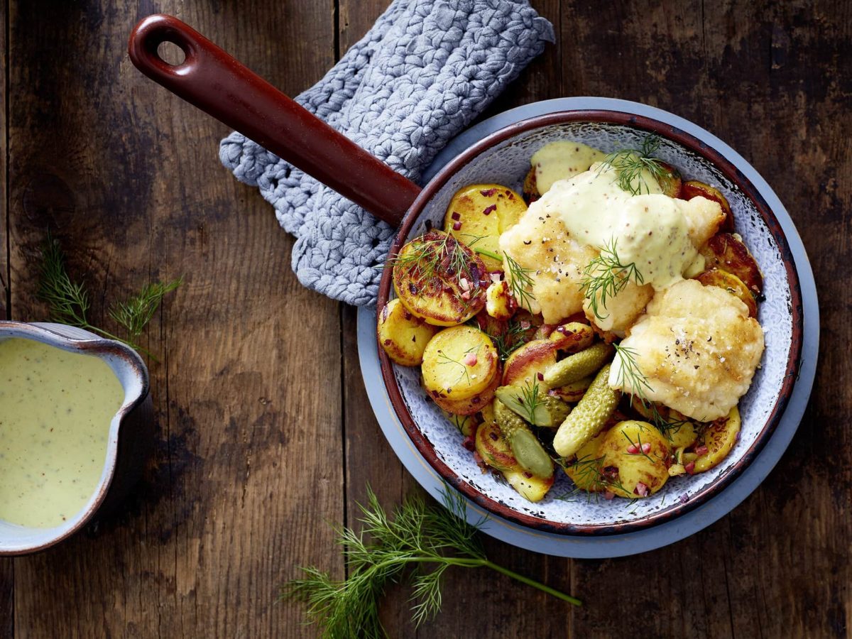 Pannfisch und Bratkartoffeln serviert in einer Pfanne. Die Senfsoße wird in einer Ausgusskanne dazu gereicht.